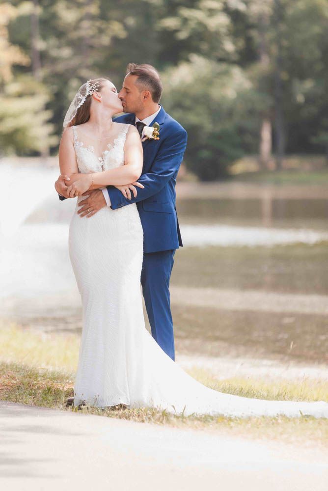 Bride in white gown and groom in navy suit share a romantic kiss outdoors near a lake on their wedding day.