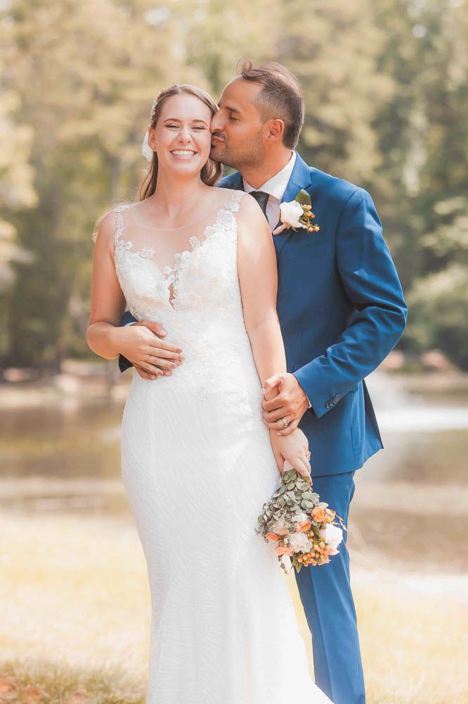 Bride in lace gown holds bouquet as groom in blue suit kisses her cheek outdoors near a sunlit pond.