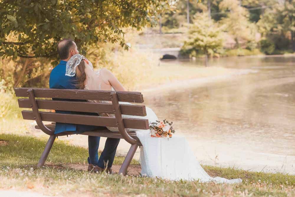 Bride and groom sitting together on a wooden bench by a serene river, surrounded by lush green trees on their wedding day.