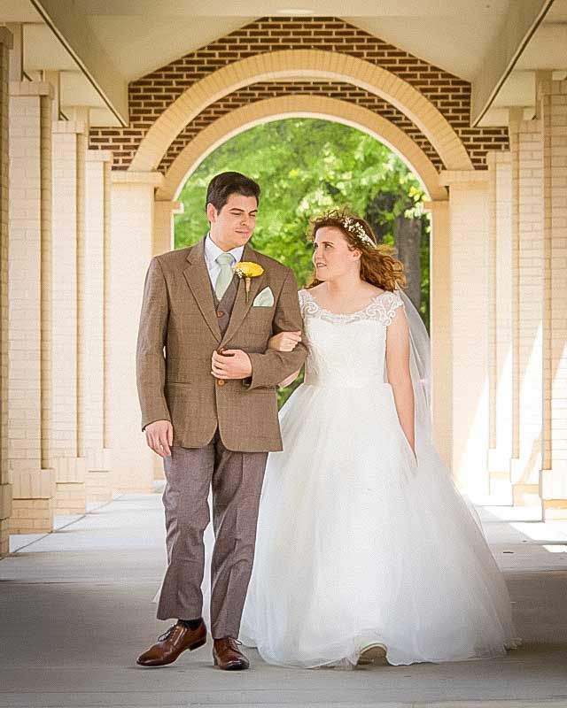 Bride and groom walking under brick arched colonnade, bride in white gown with veil, groom in tan suit with yellow boutonniere.