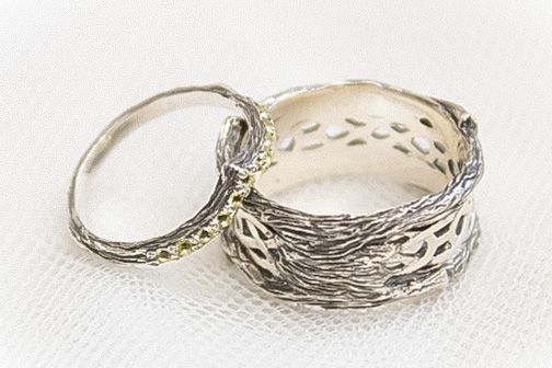 Two intricate silver wedding bands with textured bark and leaf designs rest together on a white fabric surface.