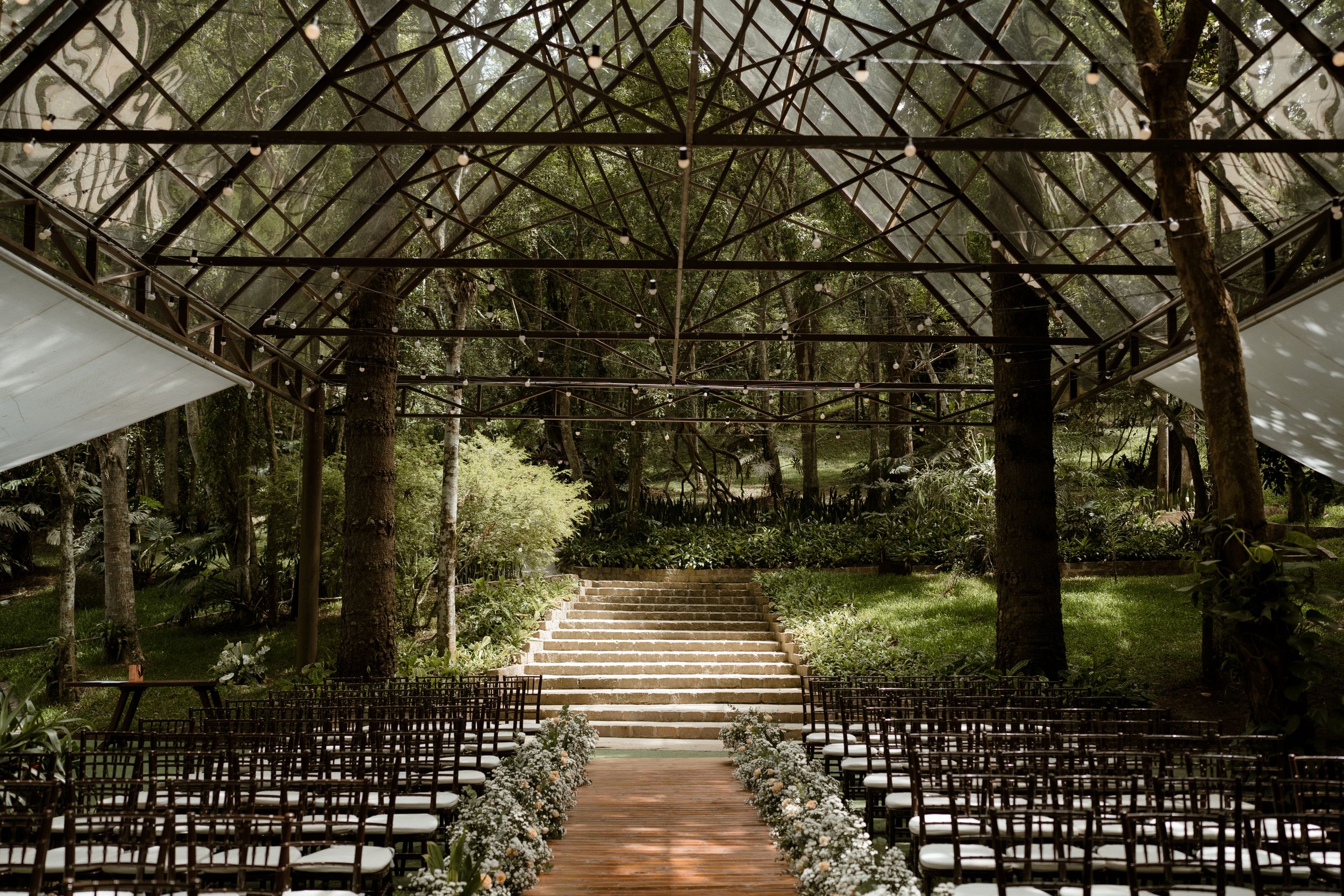 Wooden chairs line an outdoor wedding aisle beneath a forest canopy with string lights.