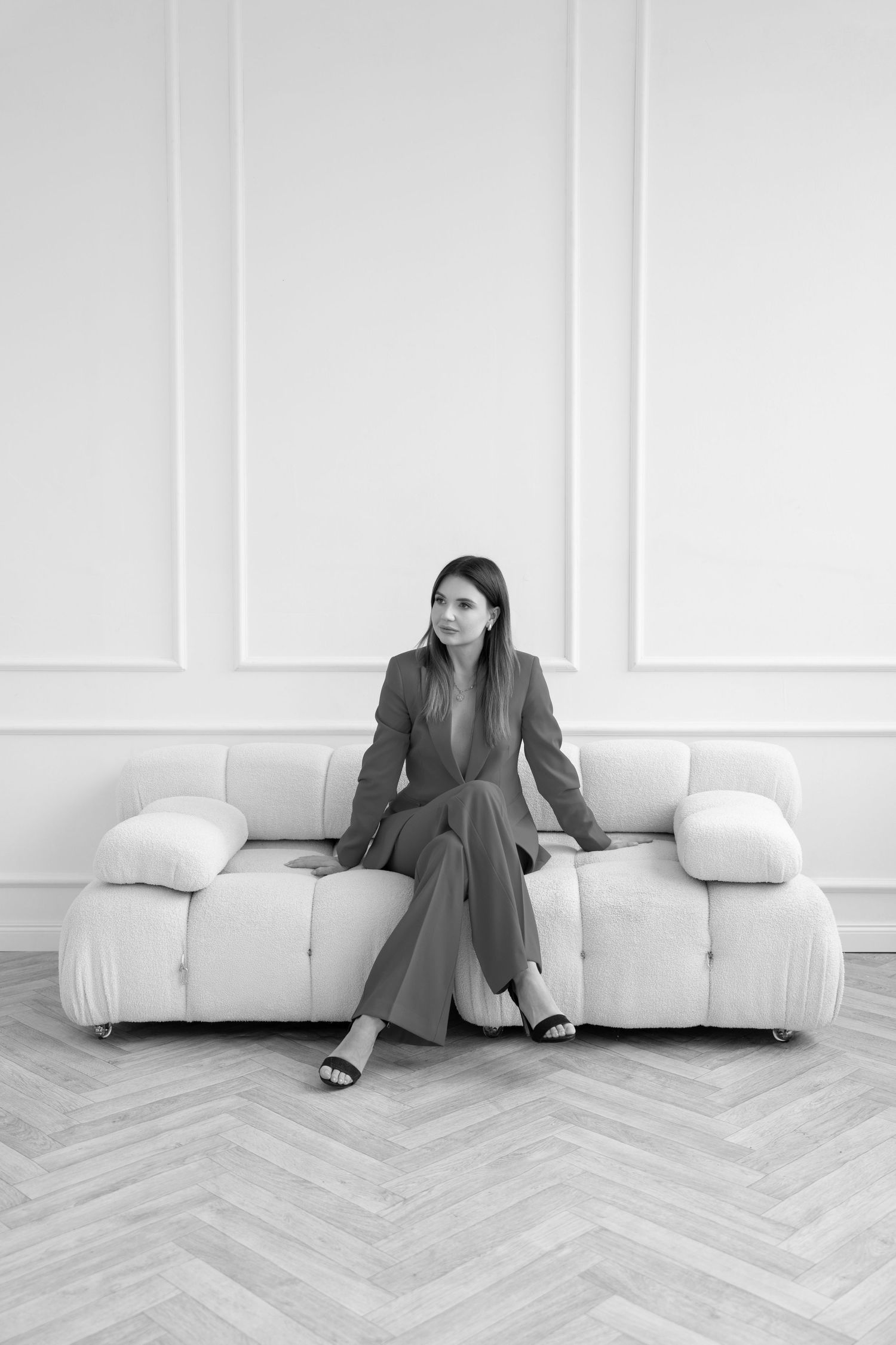Black and white photo of someone in a business suit seated alone on a white modern sofa against a minimalist white wall.