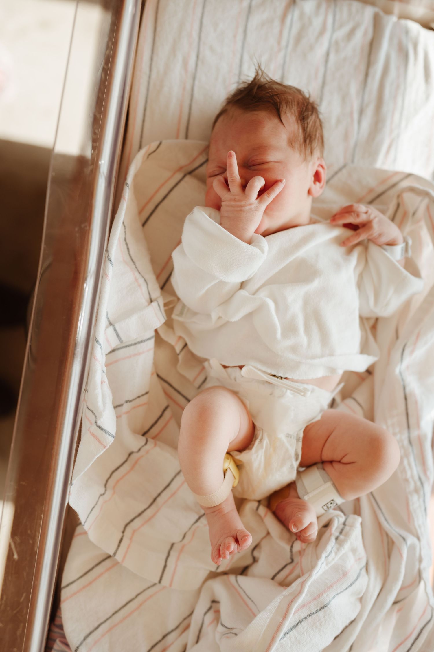 Newborn baby stretching in white clothes on cream bedding.