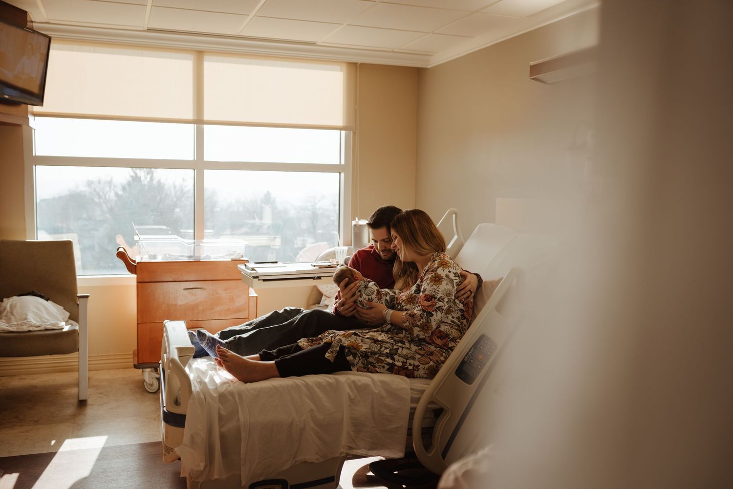 Hospital room with visitor sitting on bed near window.