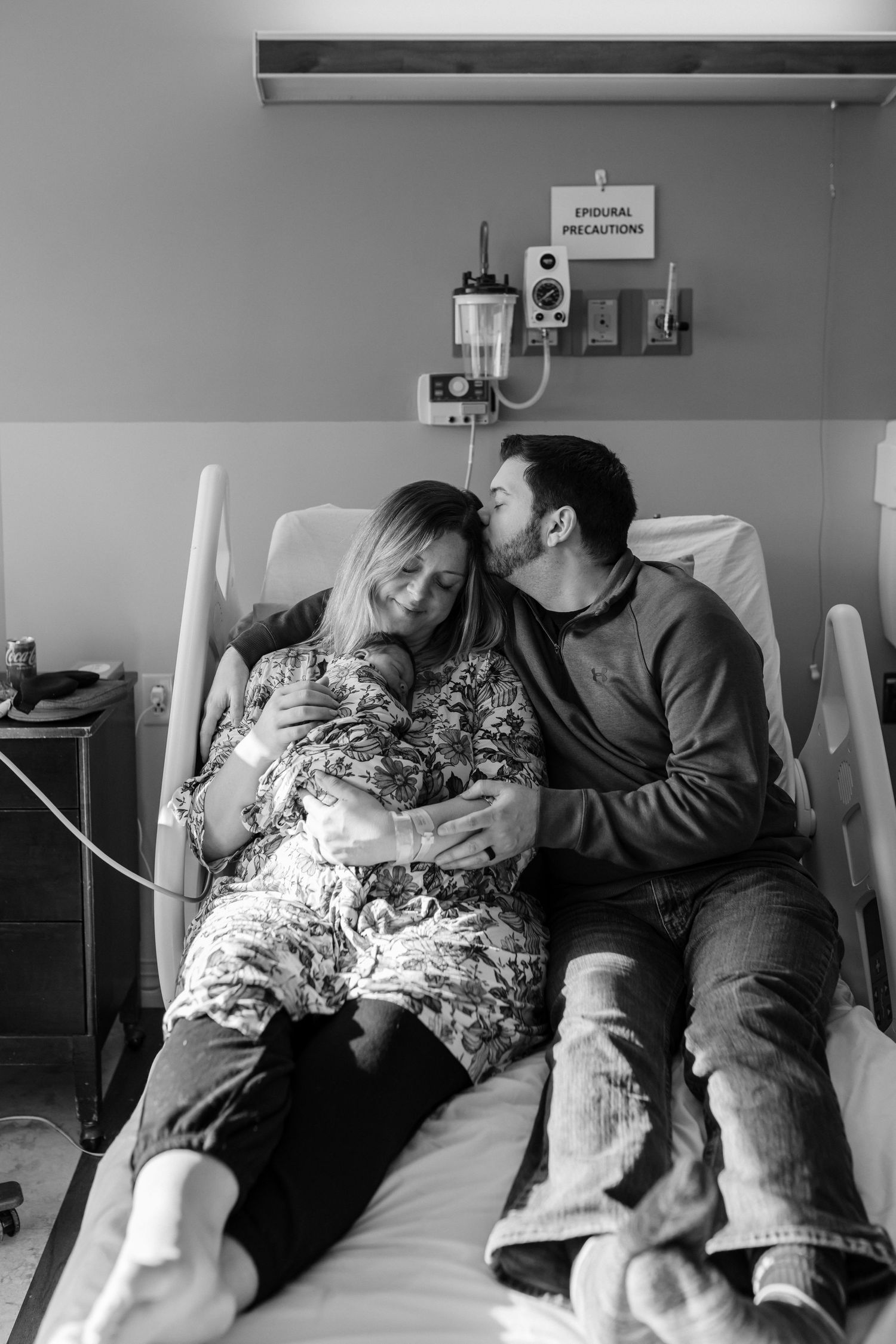 Couple shares intimate moment on hospital bed after birth, wearing casual clothes in black and white photo.