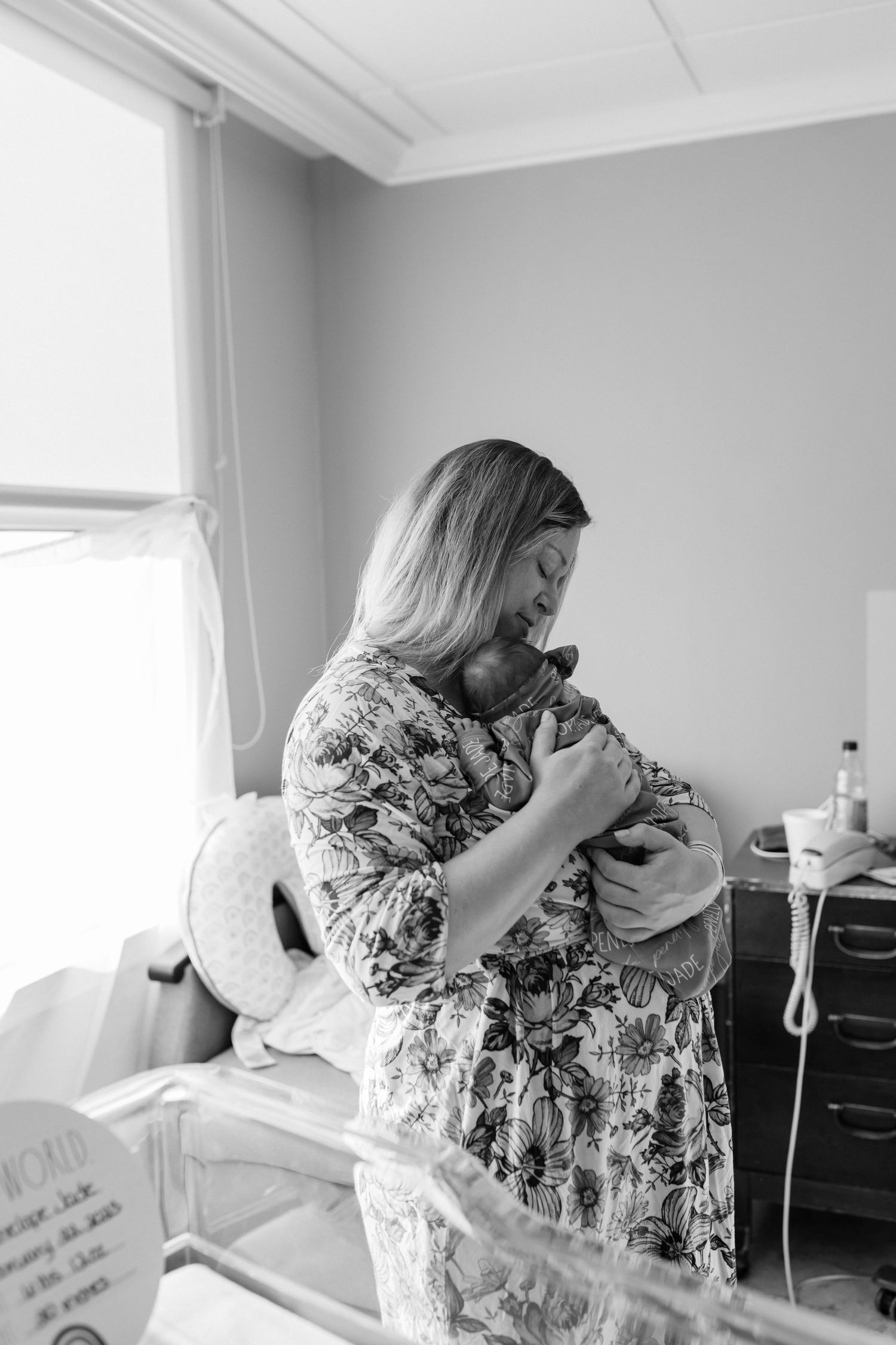 A woman in a floral dress cradles a newborn baby near a hospital window in a black and white photograph.