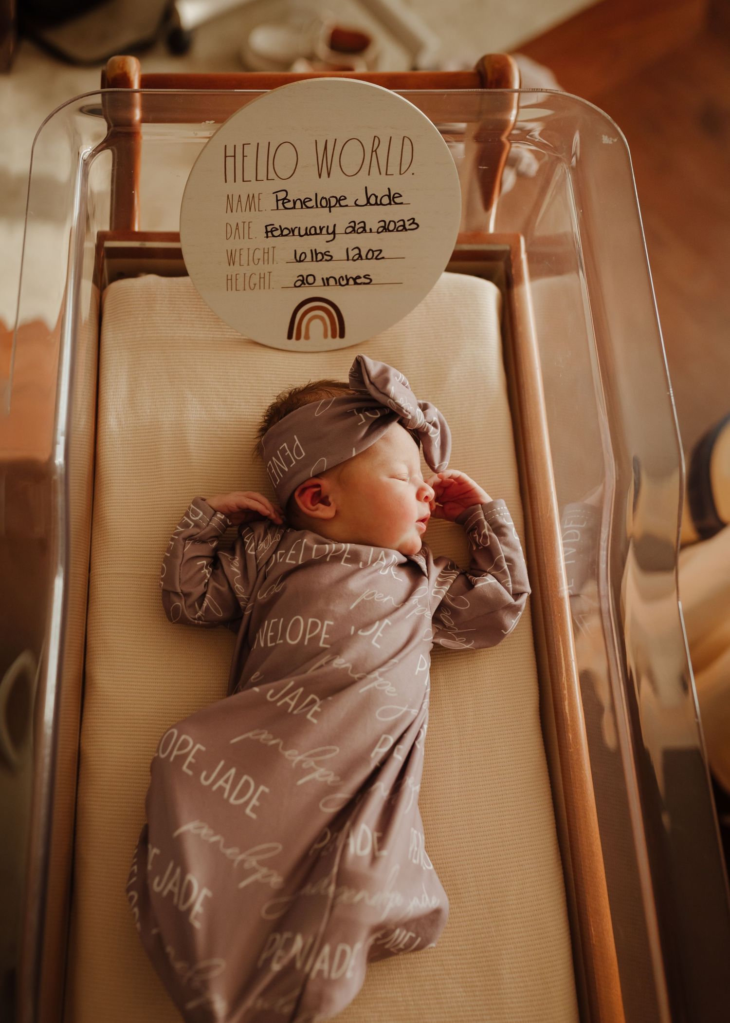 Newborn in gray onesie lying under Hello World sign in bassinet.