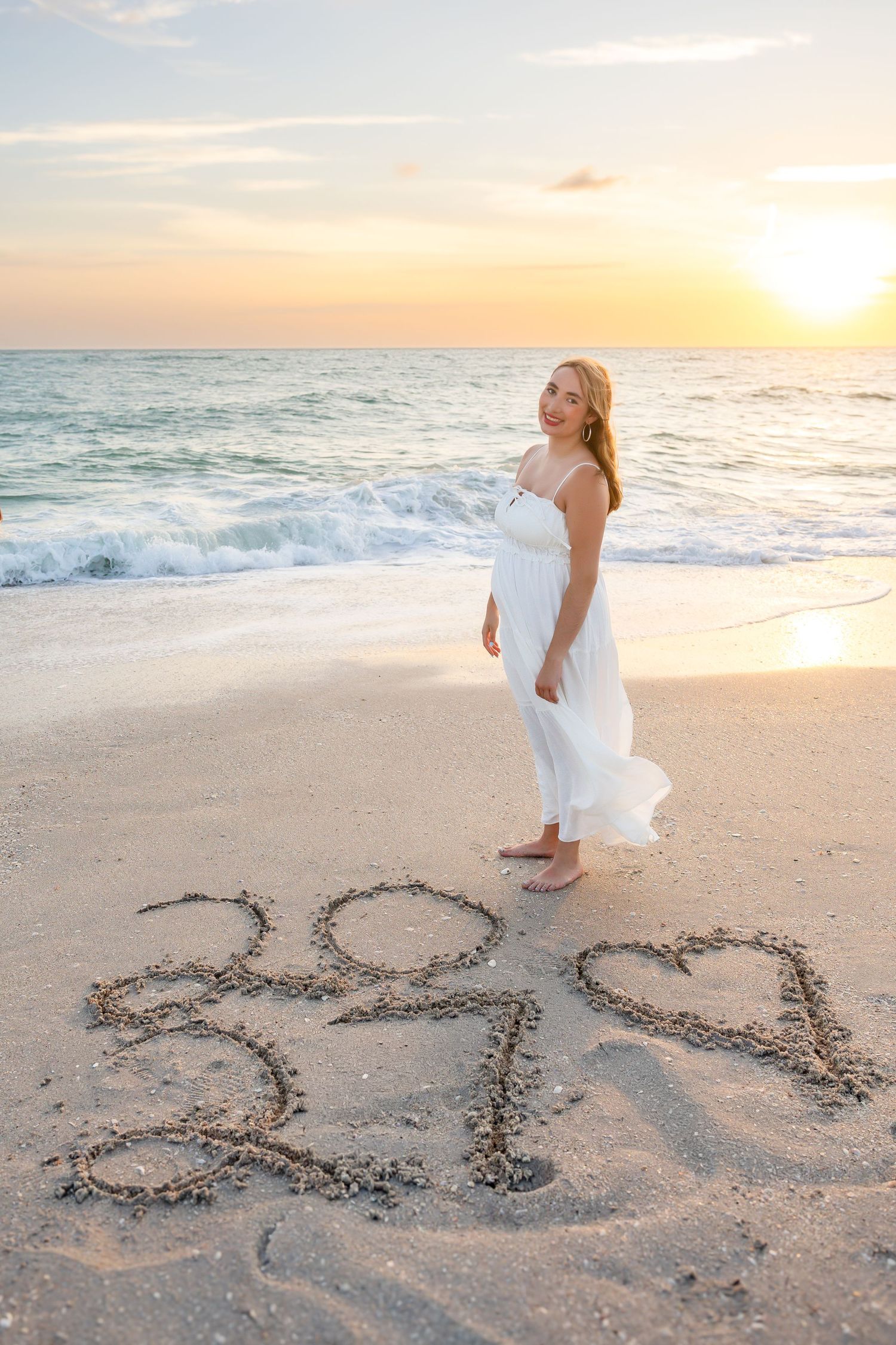 Young woman writing numbers in wet sand at beach during vibrant orange and pink sunset.