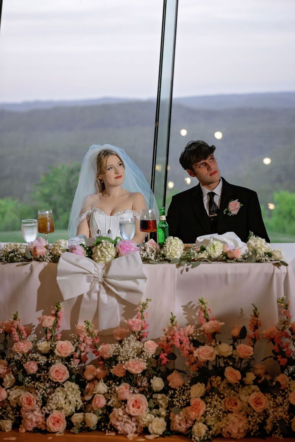 Bride and groom seated at floral-decorated sweetheart table with pink roses at elegant wedding reception.