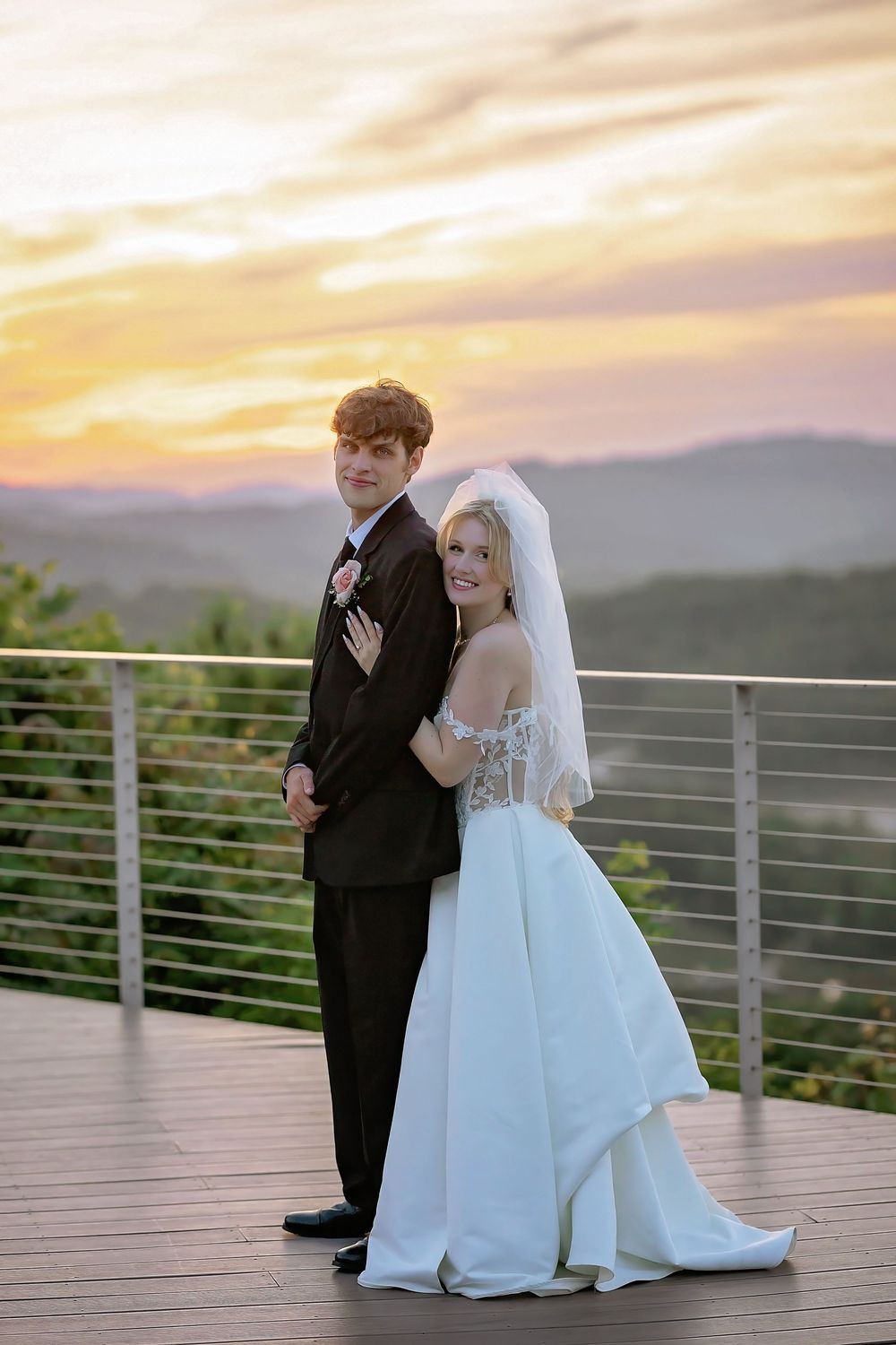 Newlywed couple posing back to back on outdoor deck with stunning mountain sunset backdrop.