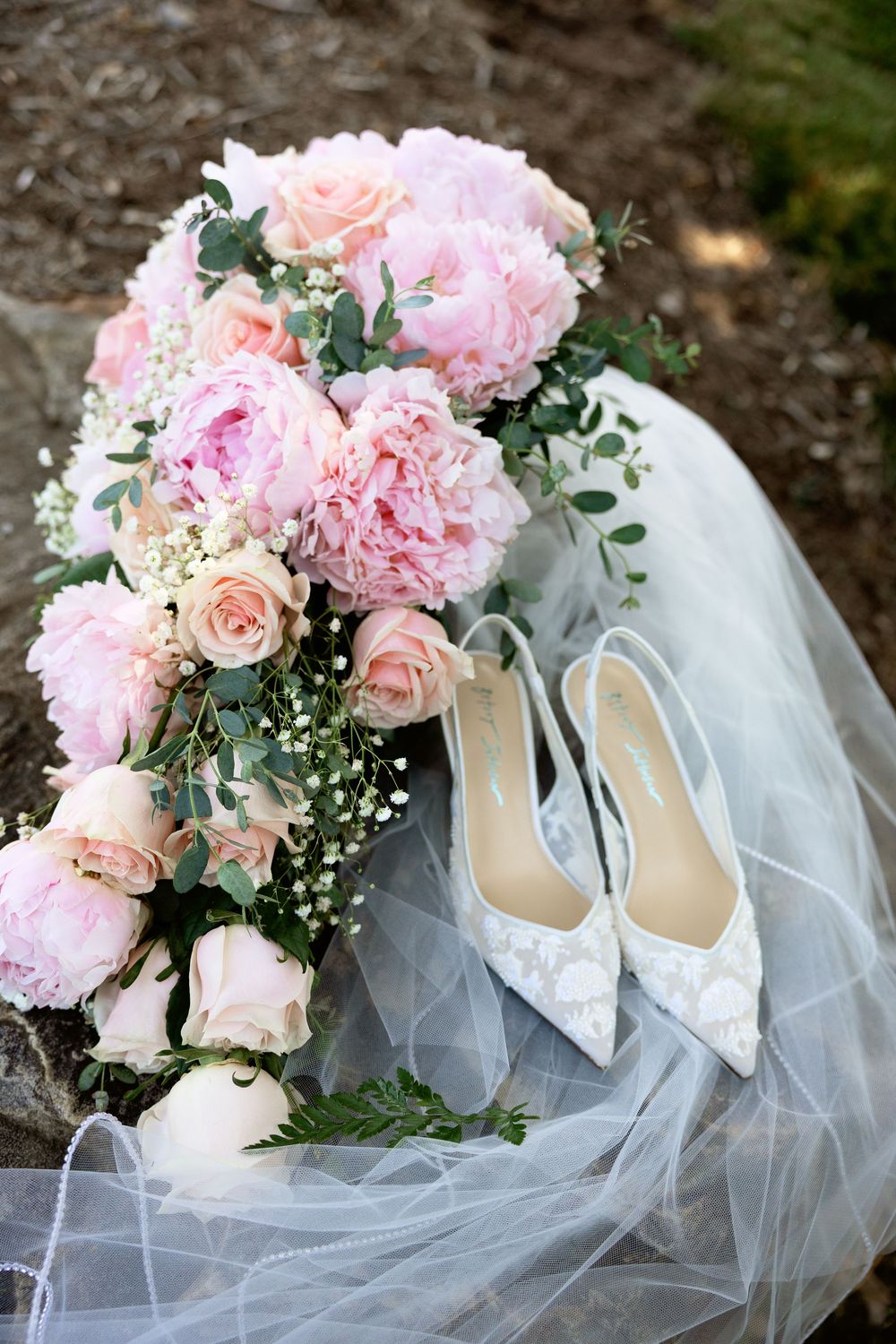 Pink peony and rose bridal bouquet beside white lace wedding shoes on tulle veil.