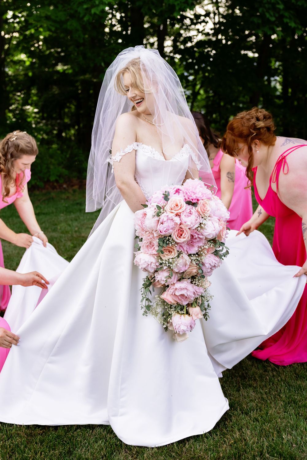 Bride in white gown holding pink floral bouquet while bridesmaids in pink dresses arrange her dress outdoors.
