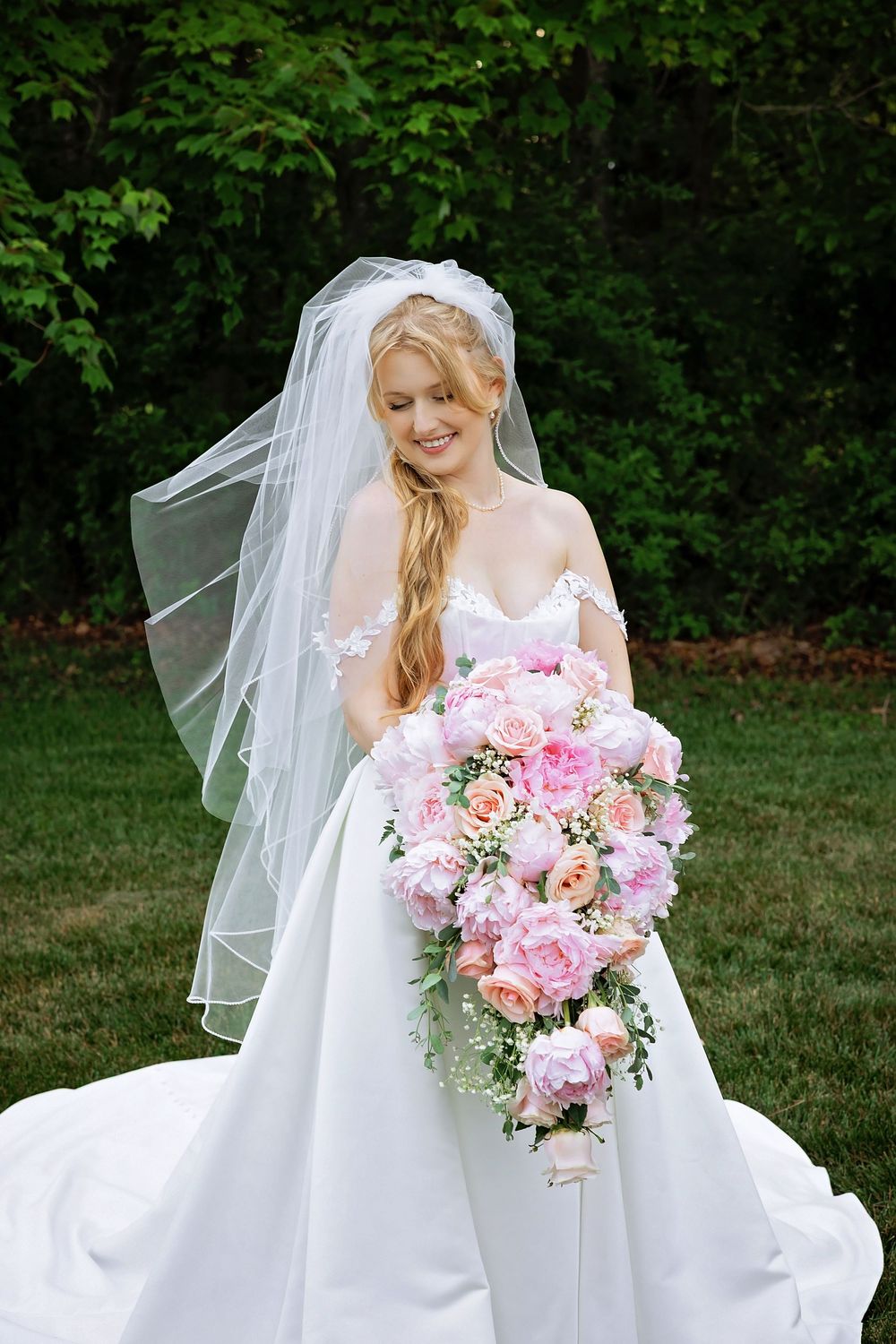 Smiling bride in white ball gown holding large cascading pink and peach bridal bouquet outdoors.