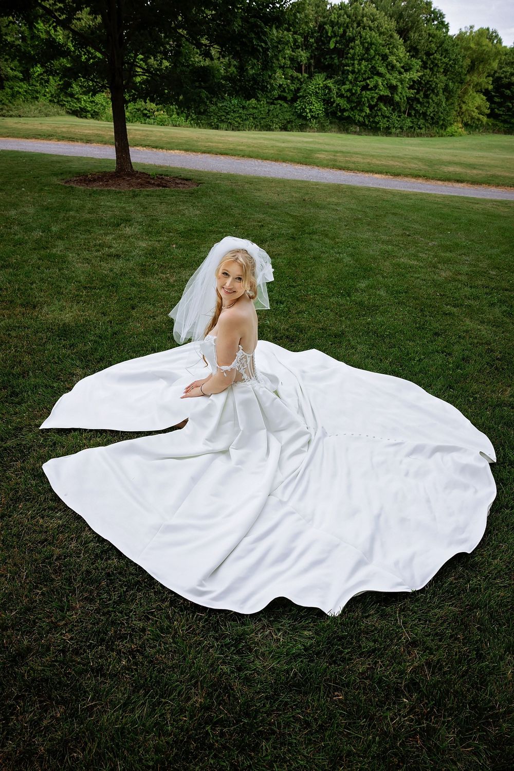 Bride in elegant white wedding gown seated on grass with dress fanned out in park.