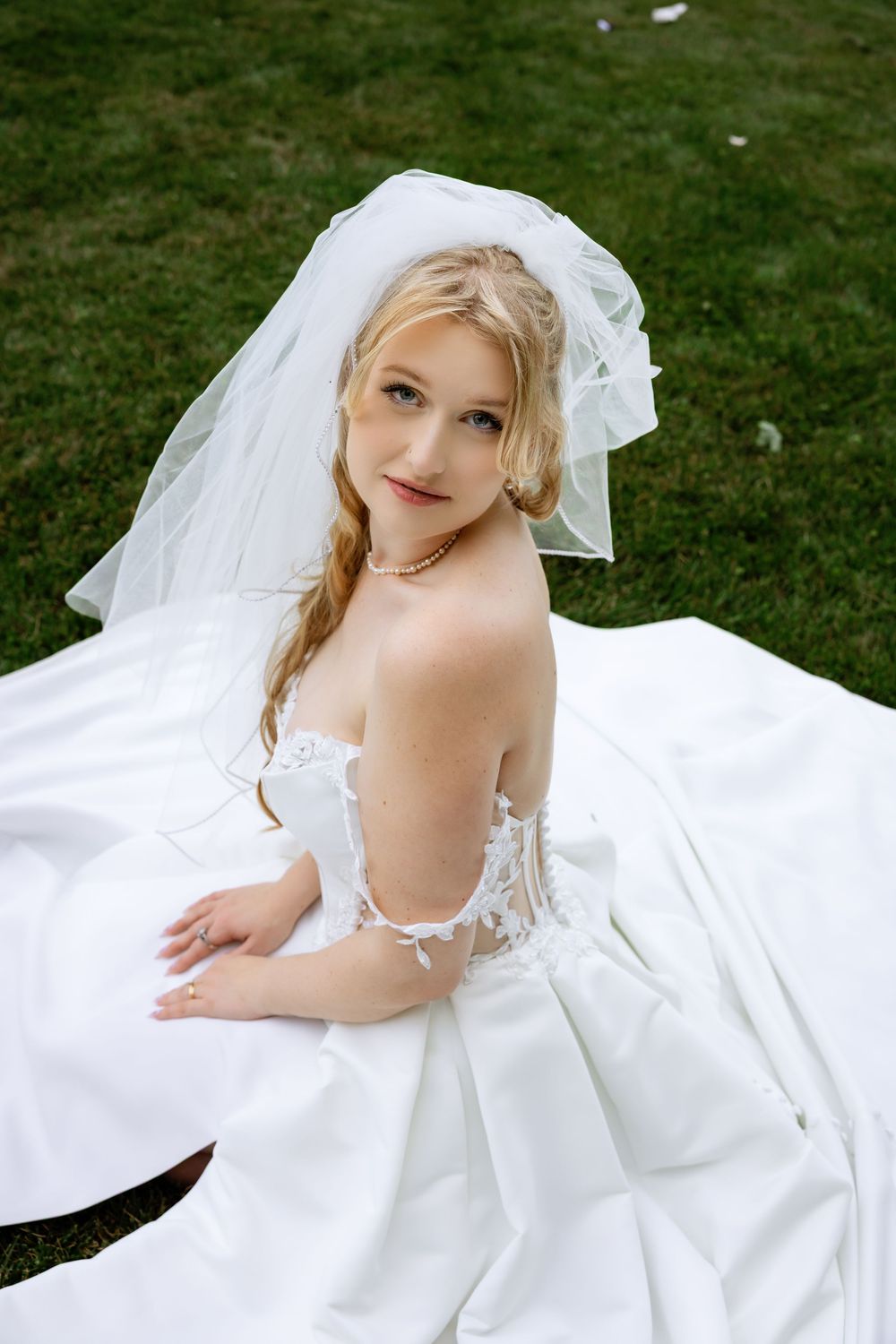 Beautiful blonde bride in white lace gown and veil sitting gracefully on green grass outdoors.