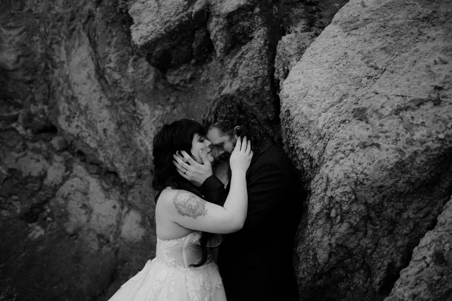 A couple embraces intimately against a rocky cliff backdrop in moody black and white lighting.