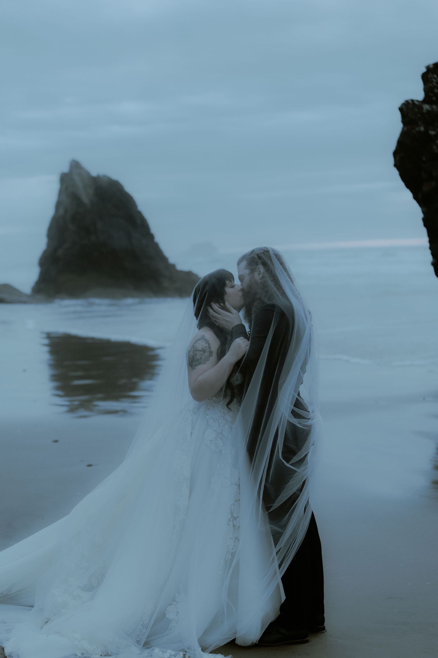 A romantic wedding portrait shows a couple embracing on a moody Pacific Northwest beach.