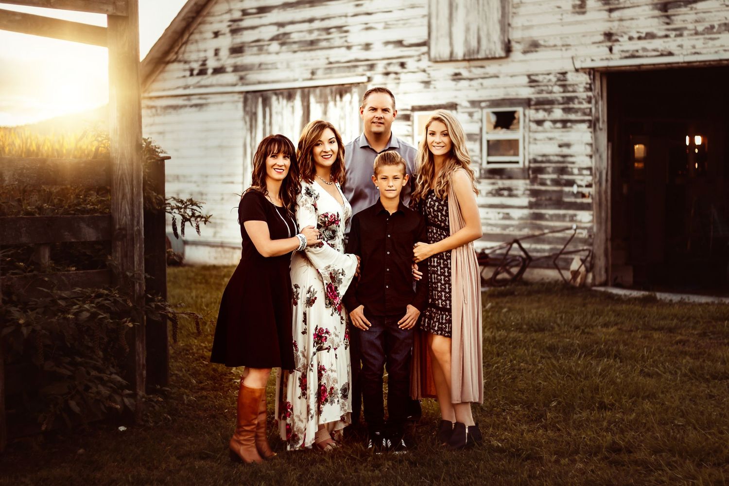Happy family of five poses together at golden hour in front of a rustic white barn in the countryside.