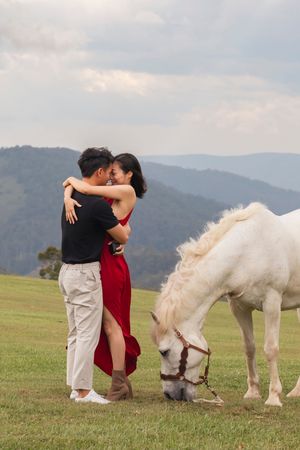 A couple shares a romantic moment near two white horses grazing in a mountain meadow.