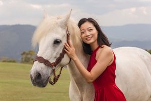Woman in flowing red dress pets gentle white horse in mountain field.