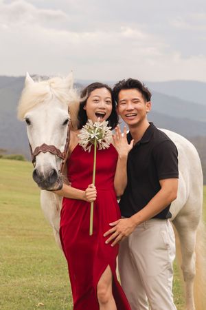A couple poses with a white horse and flowers in a scenic mountain meadow.