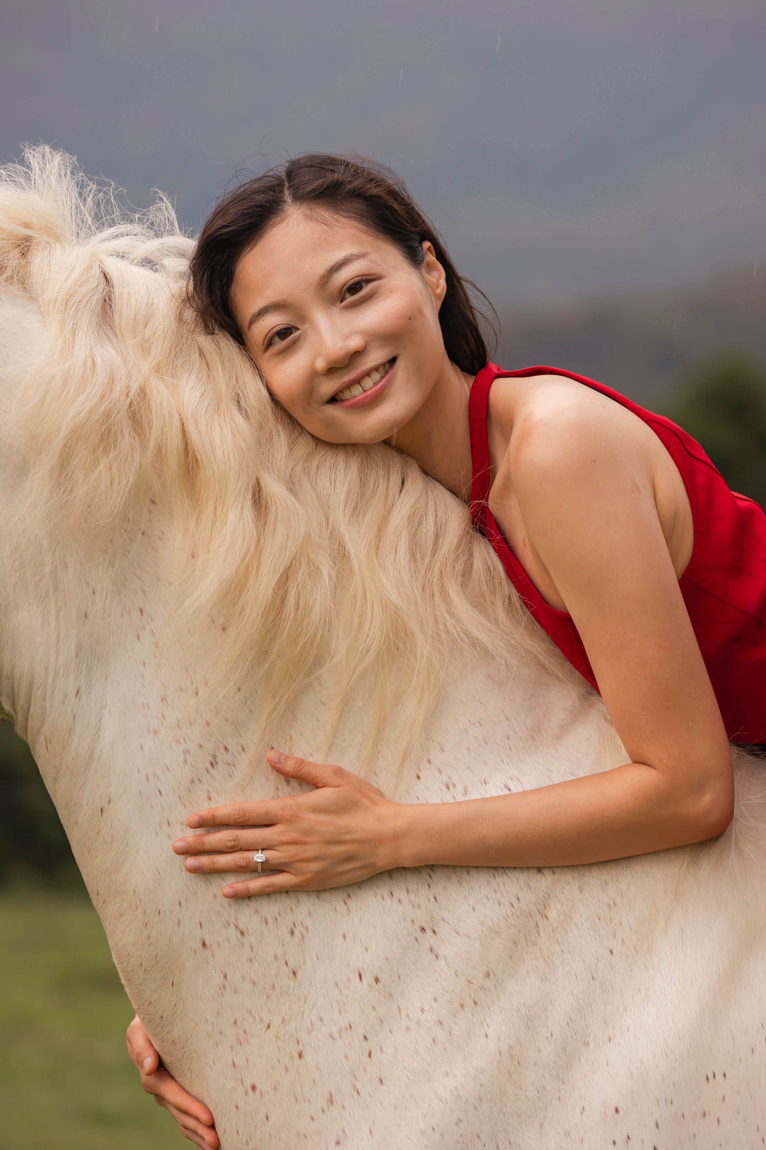 Woman in red dress hugs white horse in scenic mountain landscape.