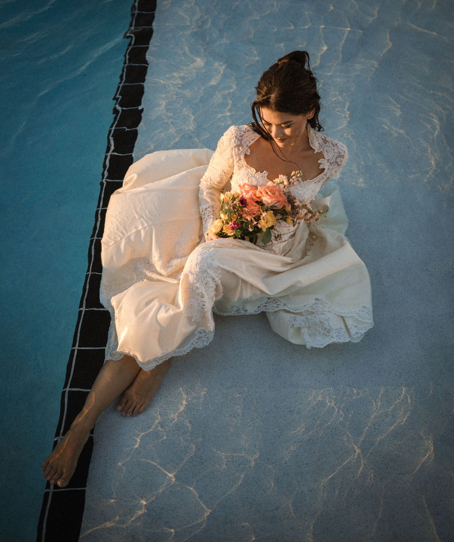 Bride in lace dress sitting poolside with colorful bouquet at sunset.