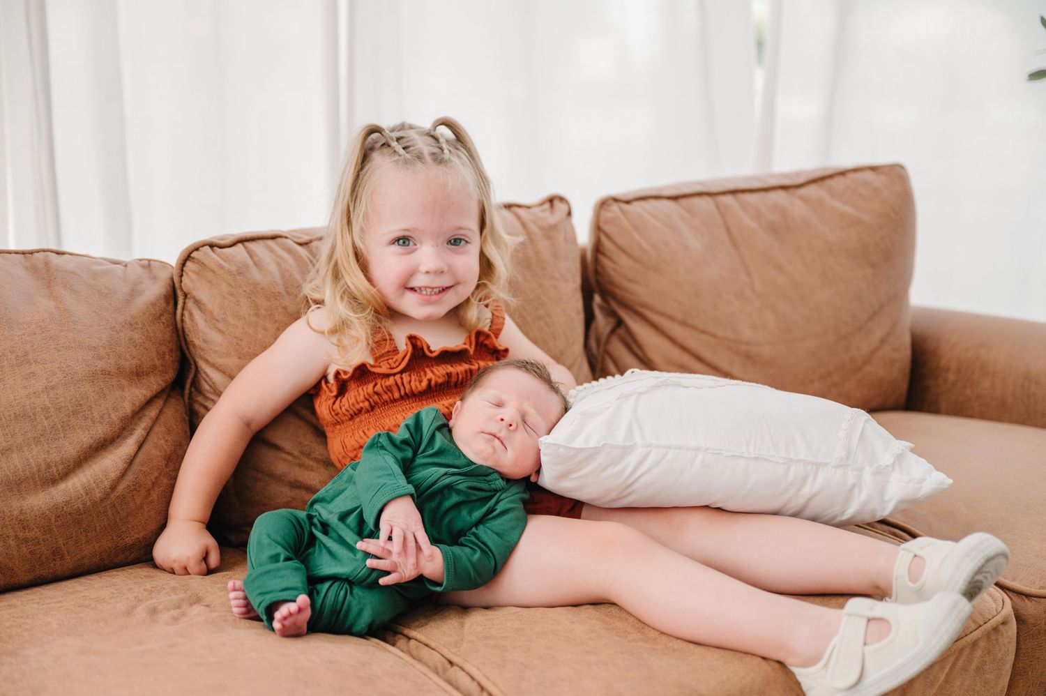 Two children relaxing together on a tan leather couch with white pillows.