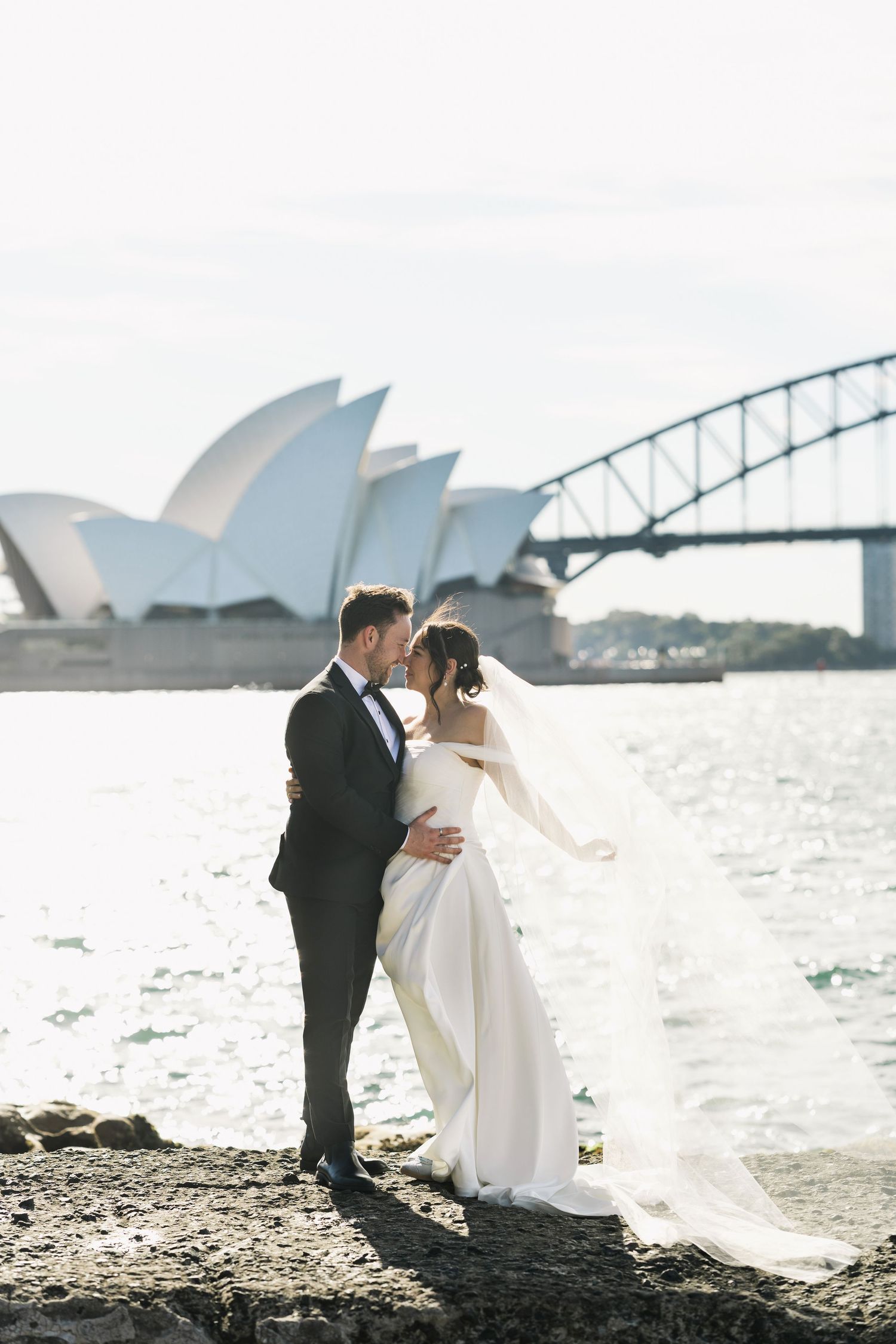 A newlywed couple standing on the rocks near Mrs Macquarie’s Chair in Sydney Botanic Garden with the Sydney Opera House in the background.