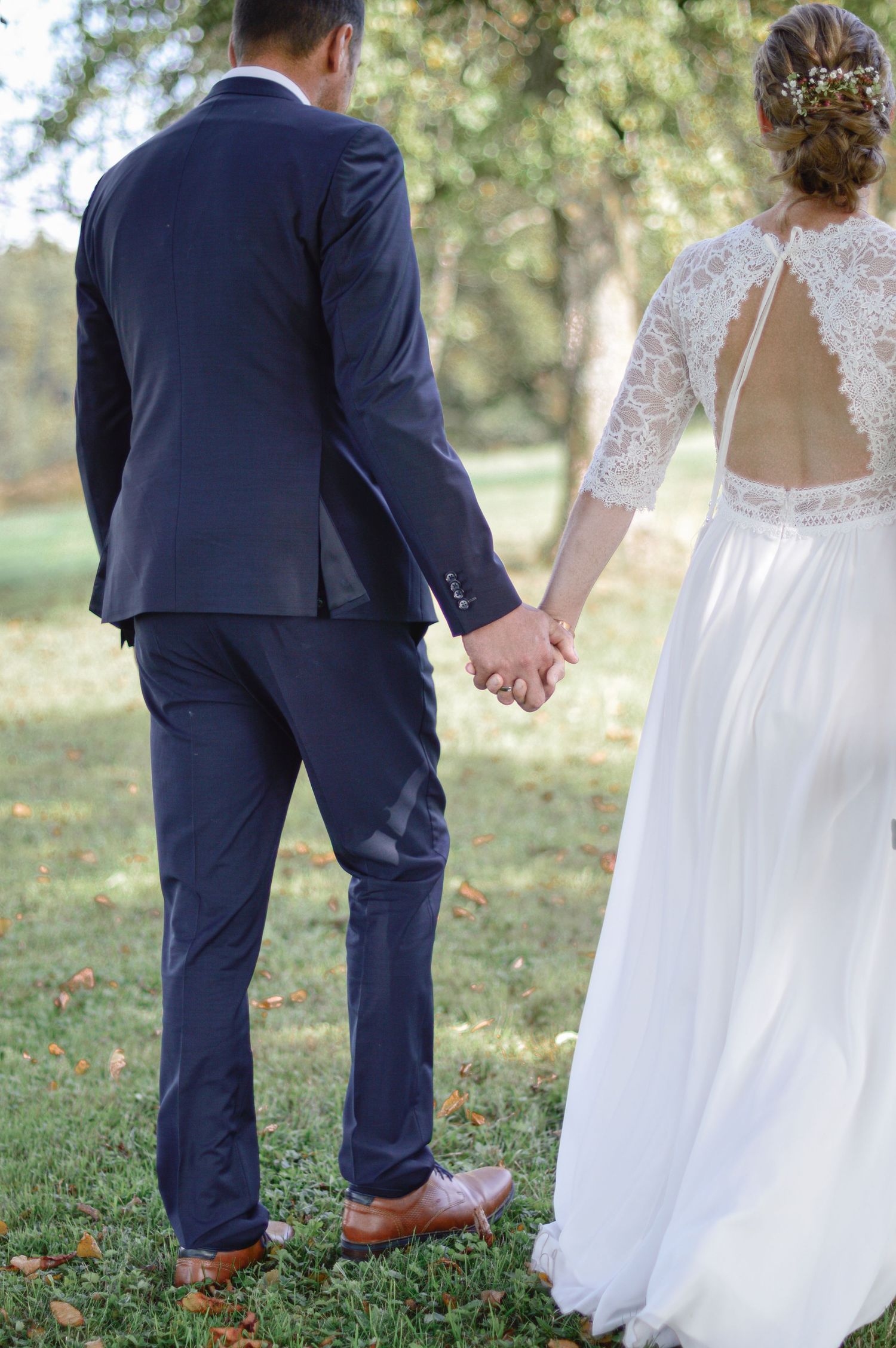 Ein Paar, das sich im Freien an den Händen hält, elegante Hochzeitskleidung trägt: ein Kleid mit Spitzenrücken und einem marineblauen Anzug.