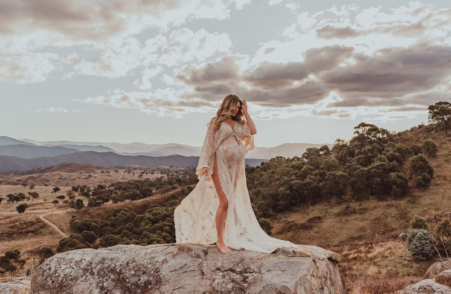 Woman in white dress standing on rocky cliff edge overlooking desert valley at sunset.