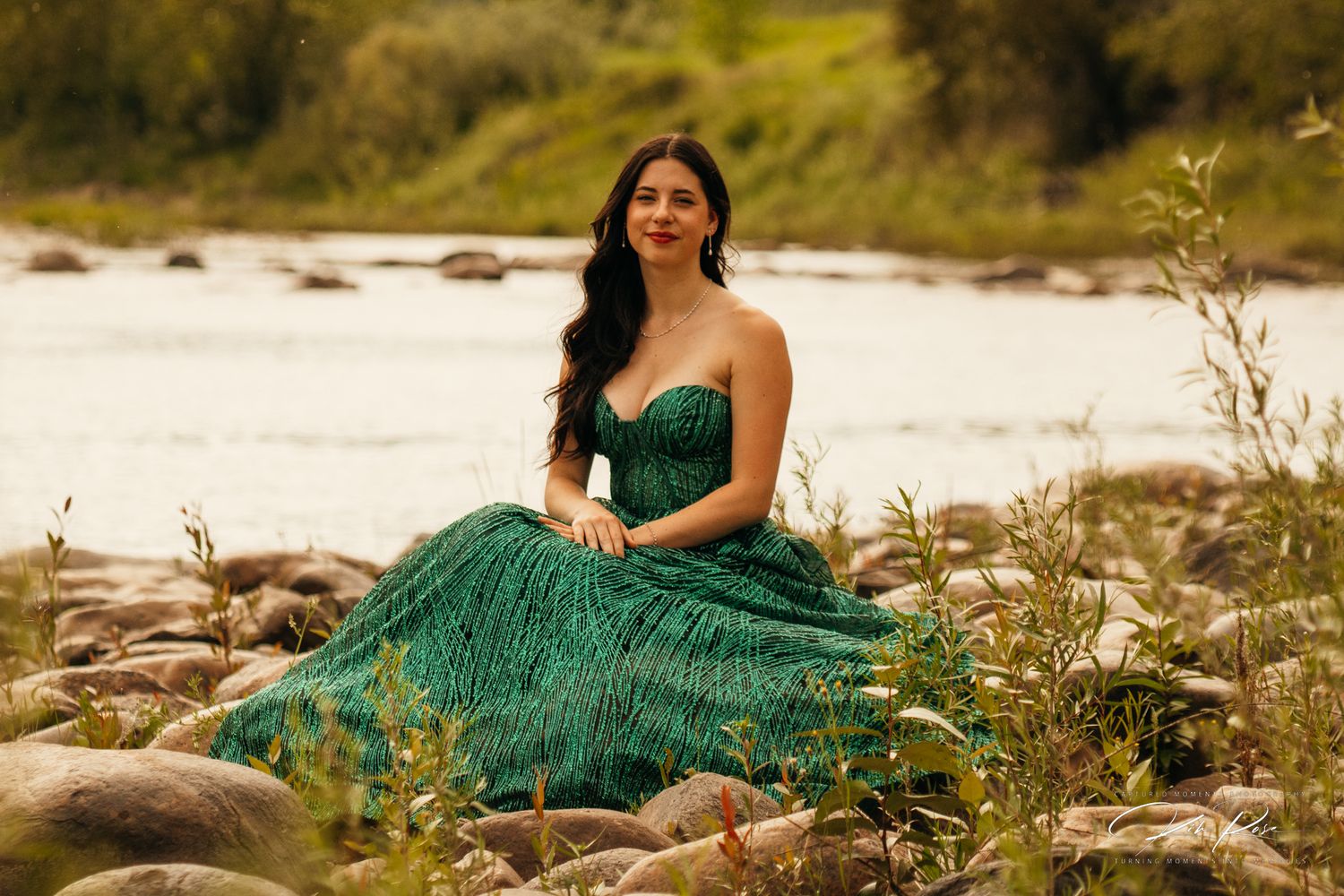 Young woman in an elegant green strapless gown sitting on rocks by a river.