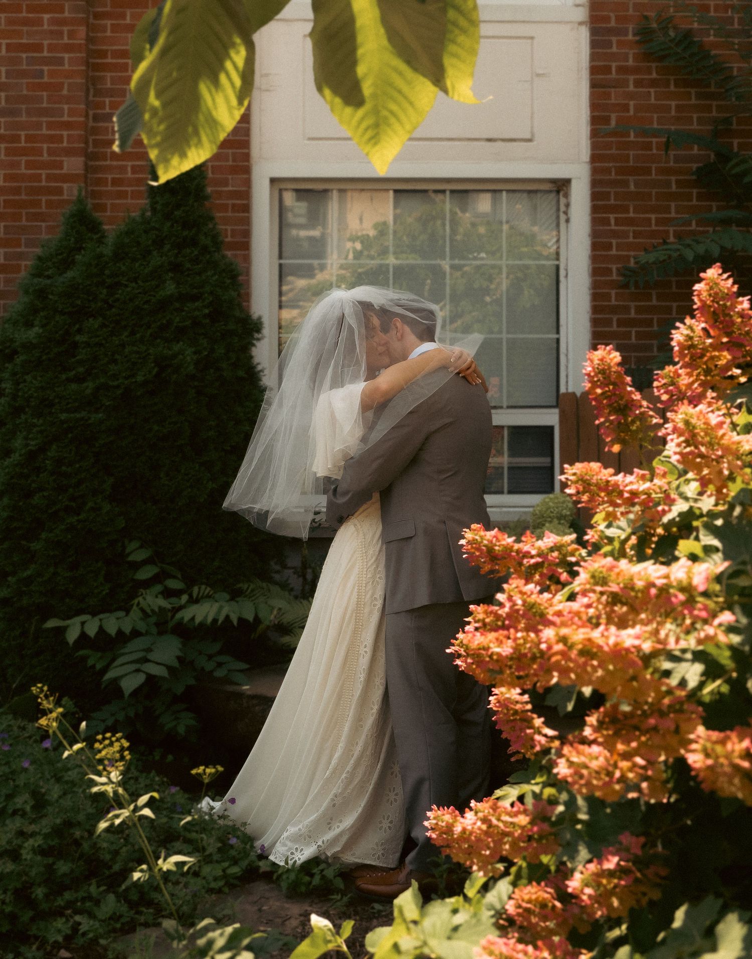 A bride and groom share an intimate embrace in a garden with orange blossoms and brick architecture.