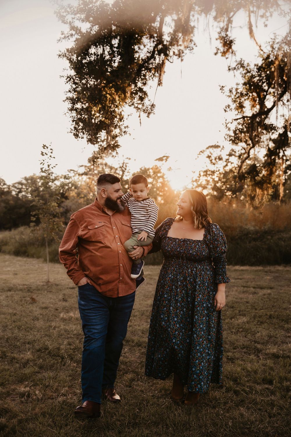 A family of three embraces in golden sunlight on grassy Houston field during sunset with trees in background.