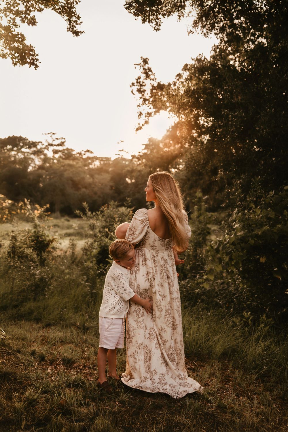 A dreamy motherhood embrace at sunset in a meadow features a floral dress and cream outfit as golden light filters through trees.