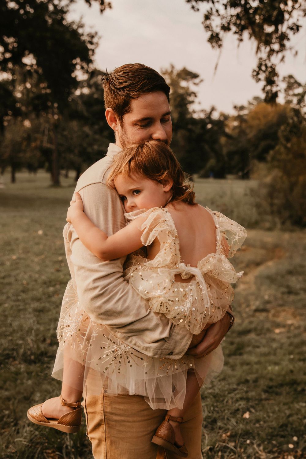 A father in a cream shirt holds a child while standing in a sunlit Houston meadow at golden hour.