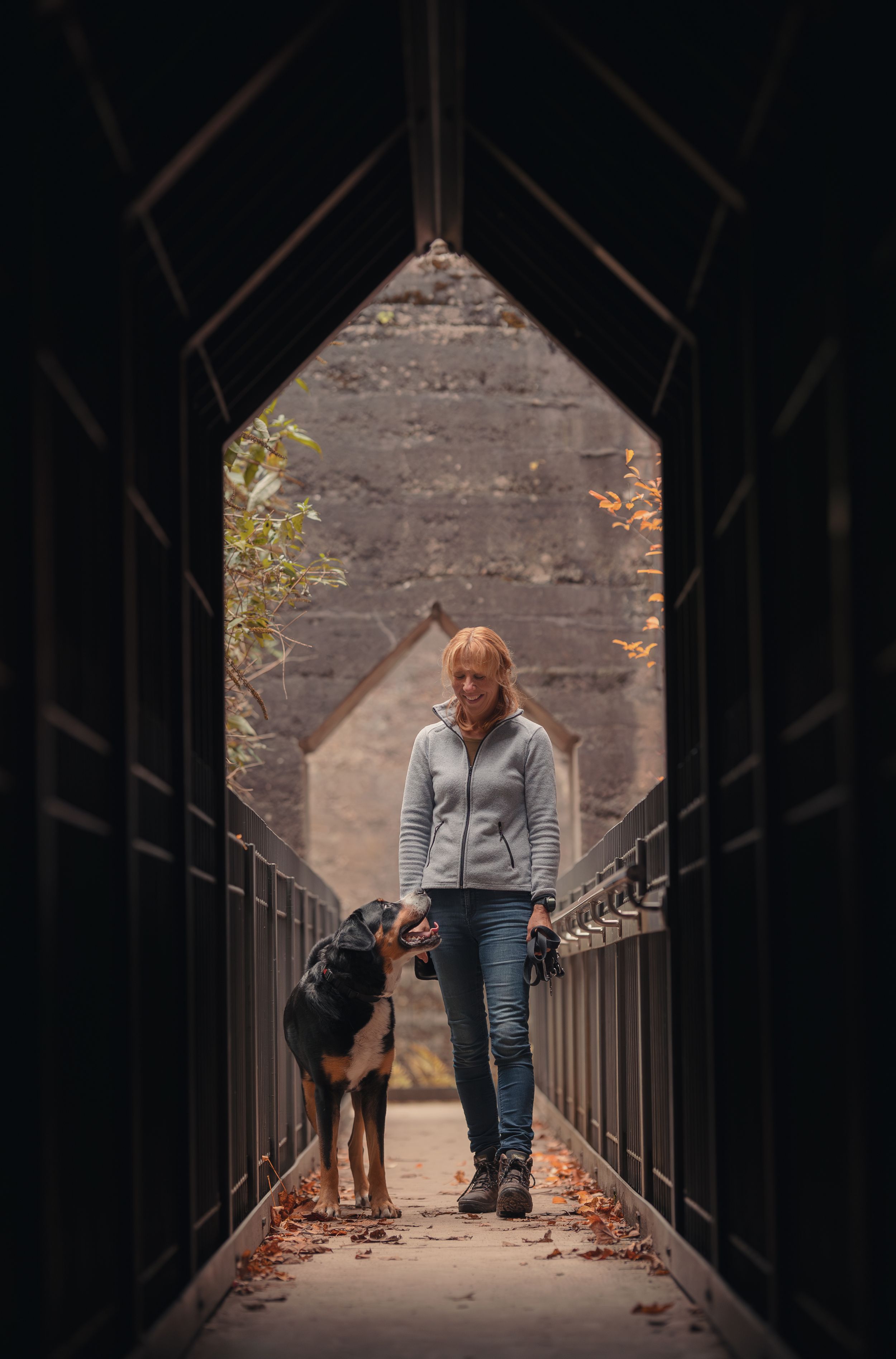 A person and their dog walk together on a narrow bridge through a dark tunnel.