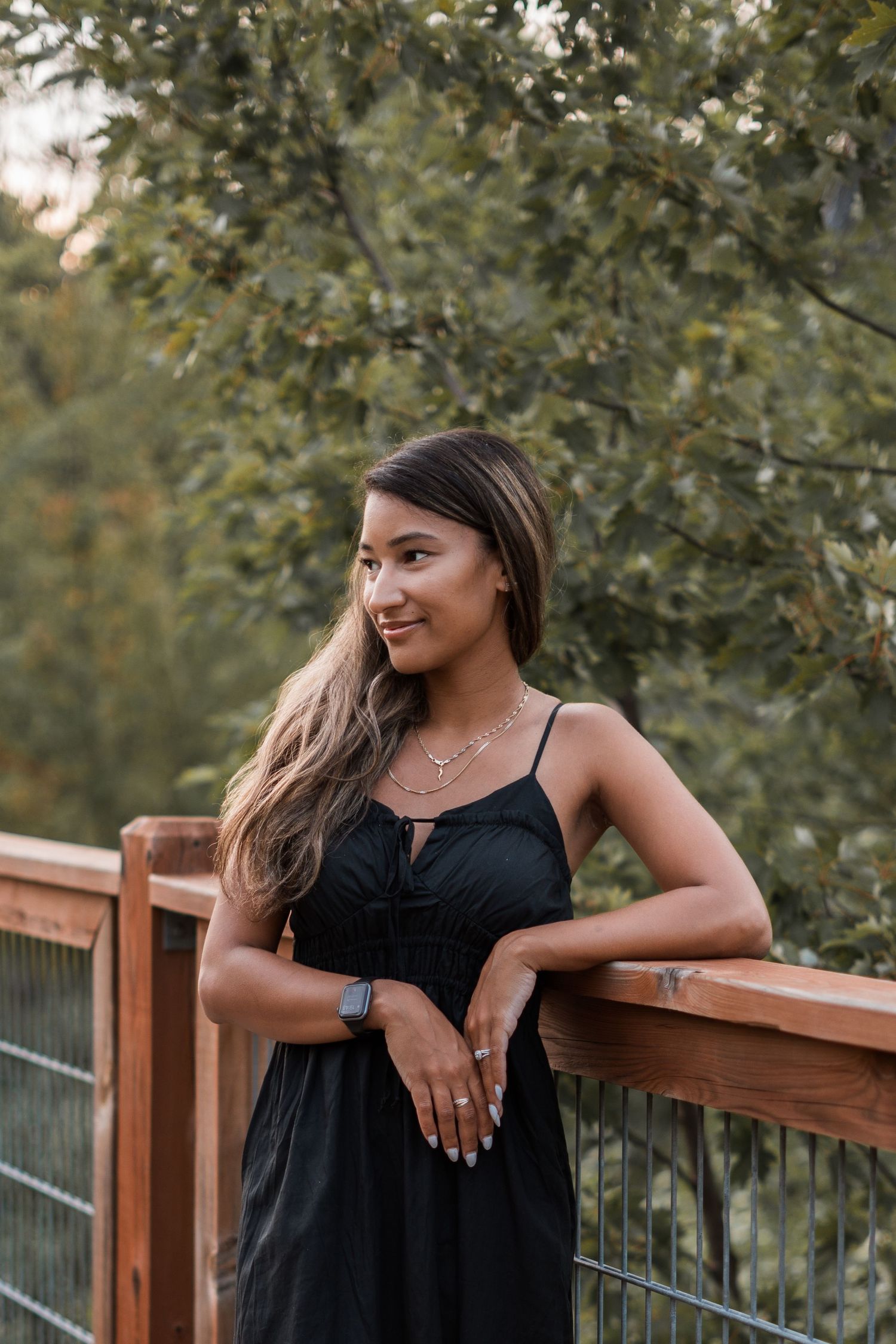 Woman in black dress smiles while leaning on wooden deck railing surrounded by green trees.