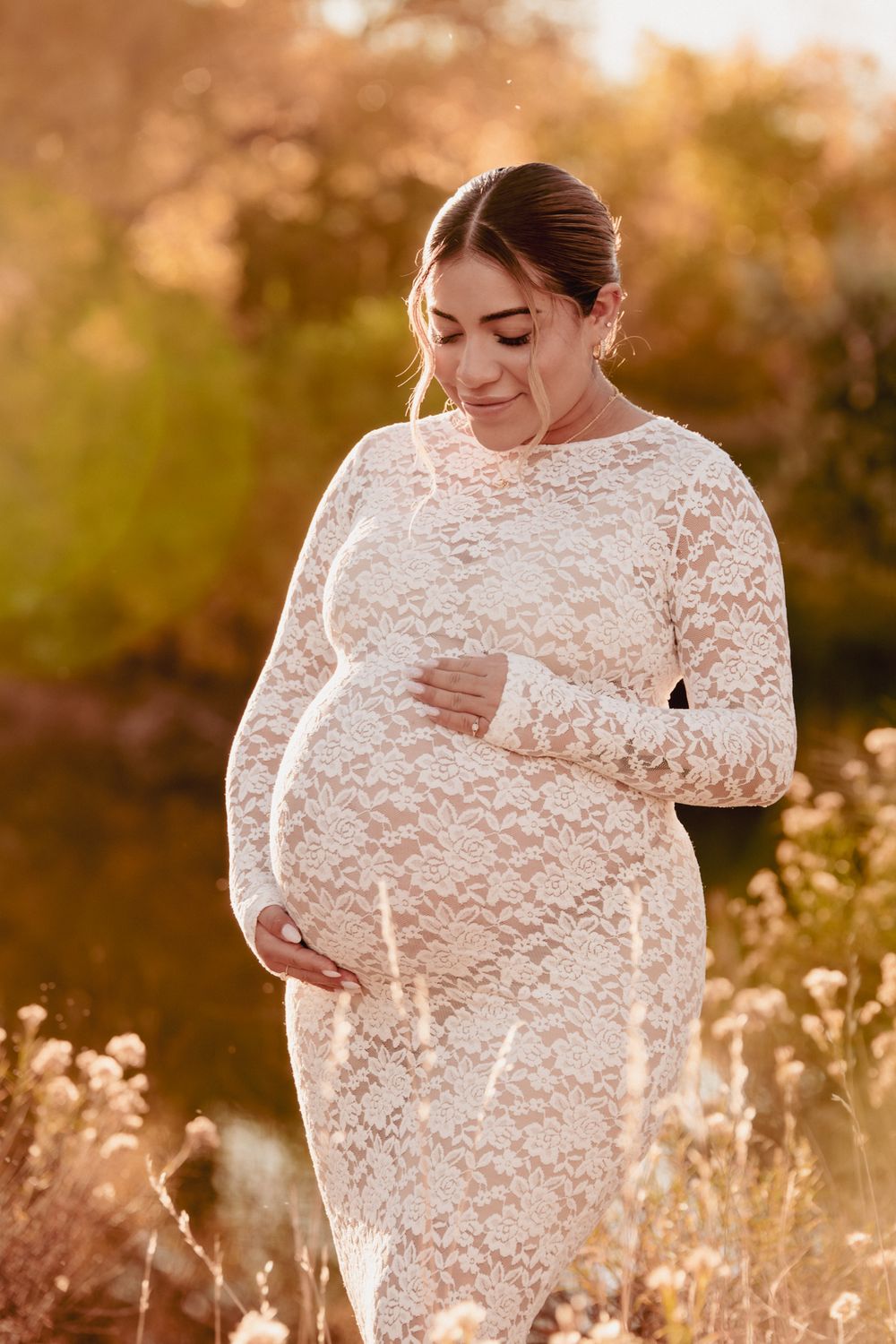 Expectant mother in white lace dress cradles baby bump during golden hour photoshoot in wildflower field.