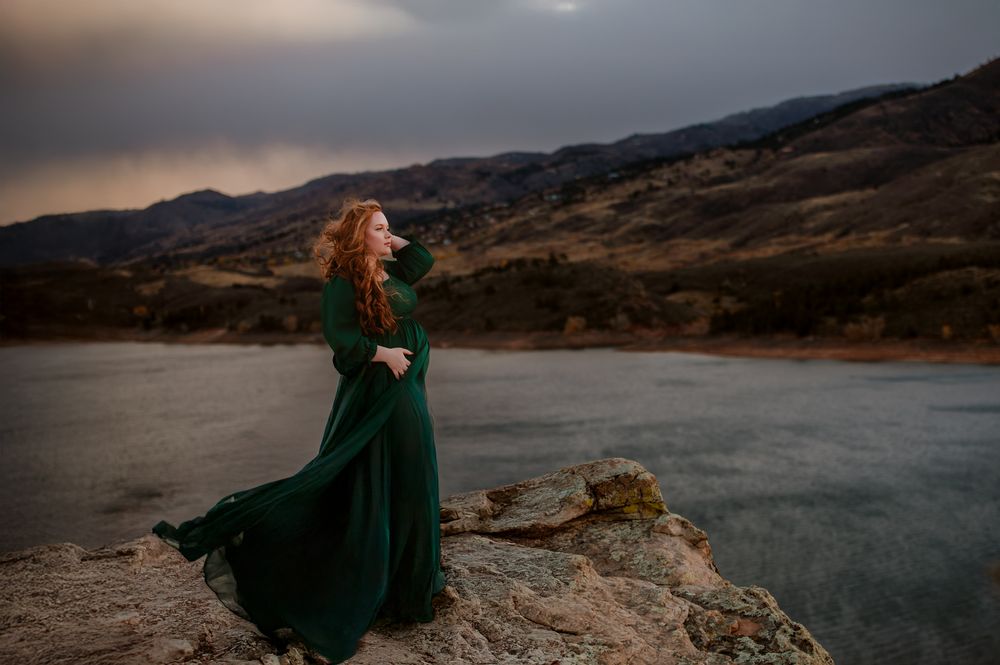 A woman in an emerald green dress poses gracefully on rocky cliffs overlooking a misty mountain lake at sunset.