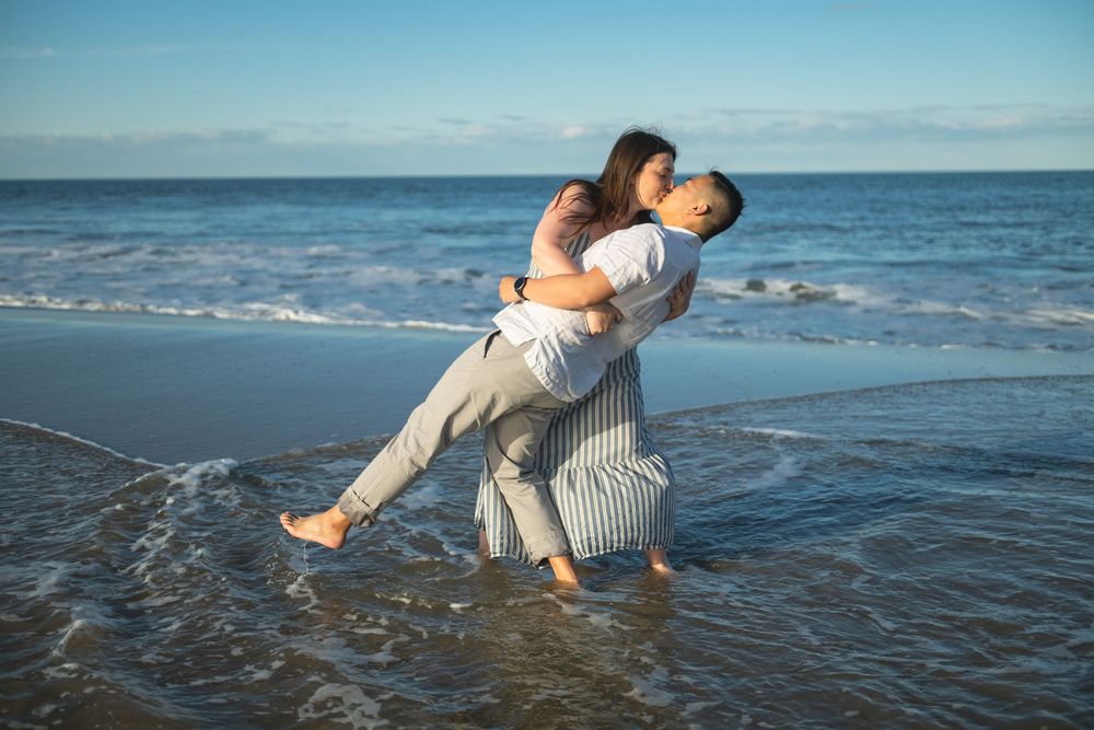 Couple embracing romantically on a sunny beach, man lifting woman in shallow ocean waves with blue sky backdrop.