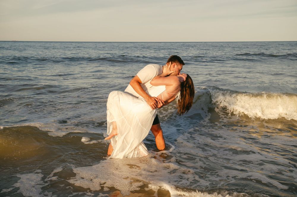 Couple sharing a romantic kiss while standing in ocean waves at sunset, bride wearing a white wedding dress.