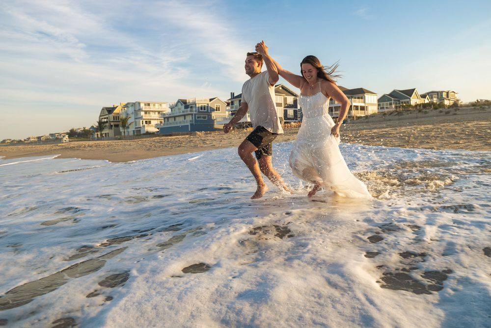 Joyful couple running through ocean waves on a sandy beach at sunset, bride in white gown, groom in casual attire.