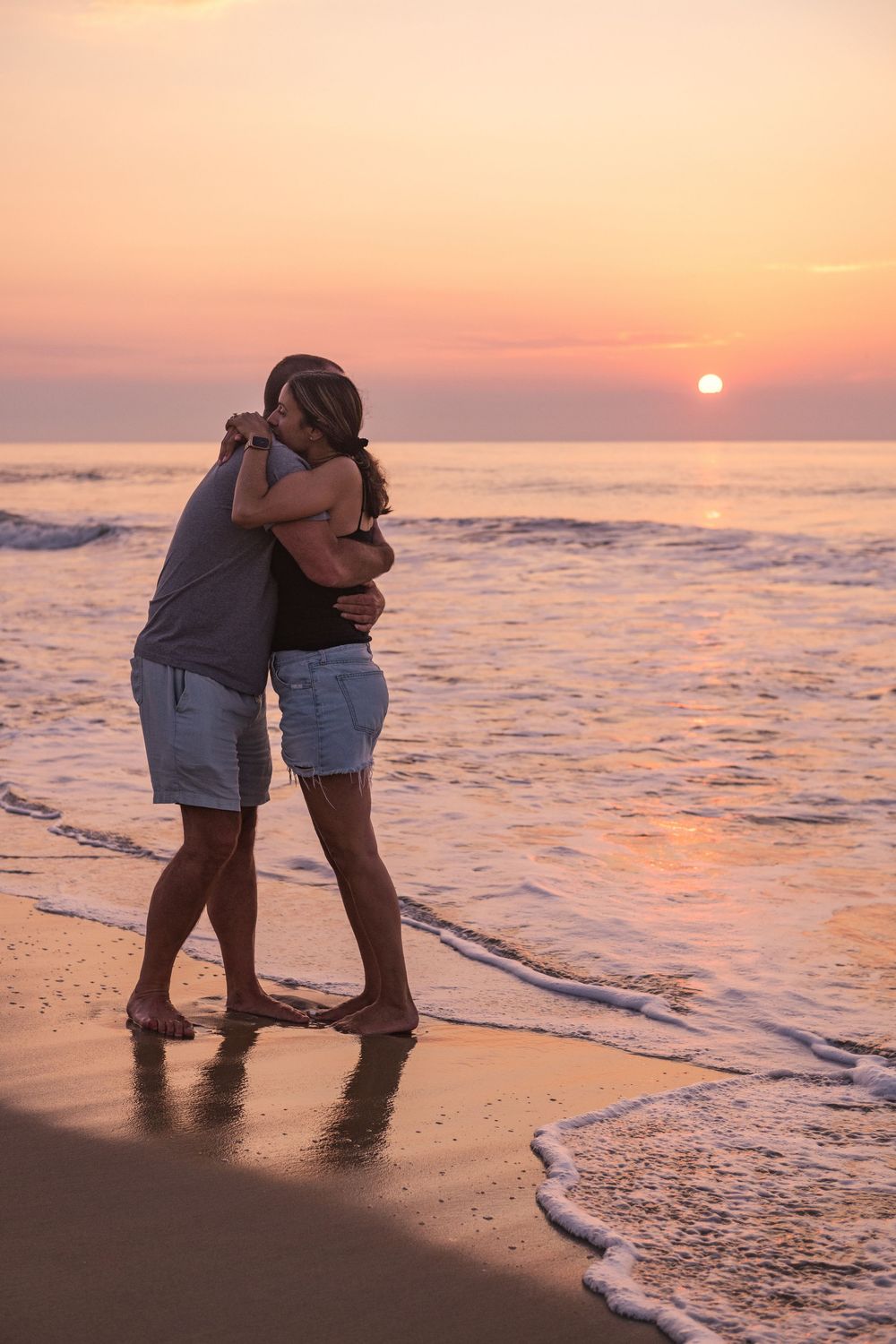 A couple embracing on the beach at sunset with ocean waves lapping at their feet during golden hour.