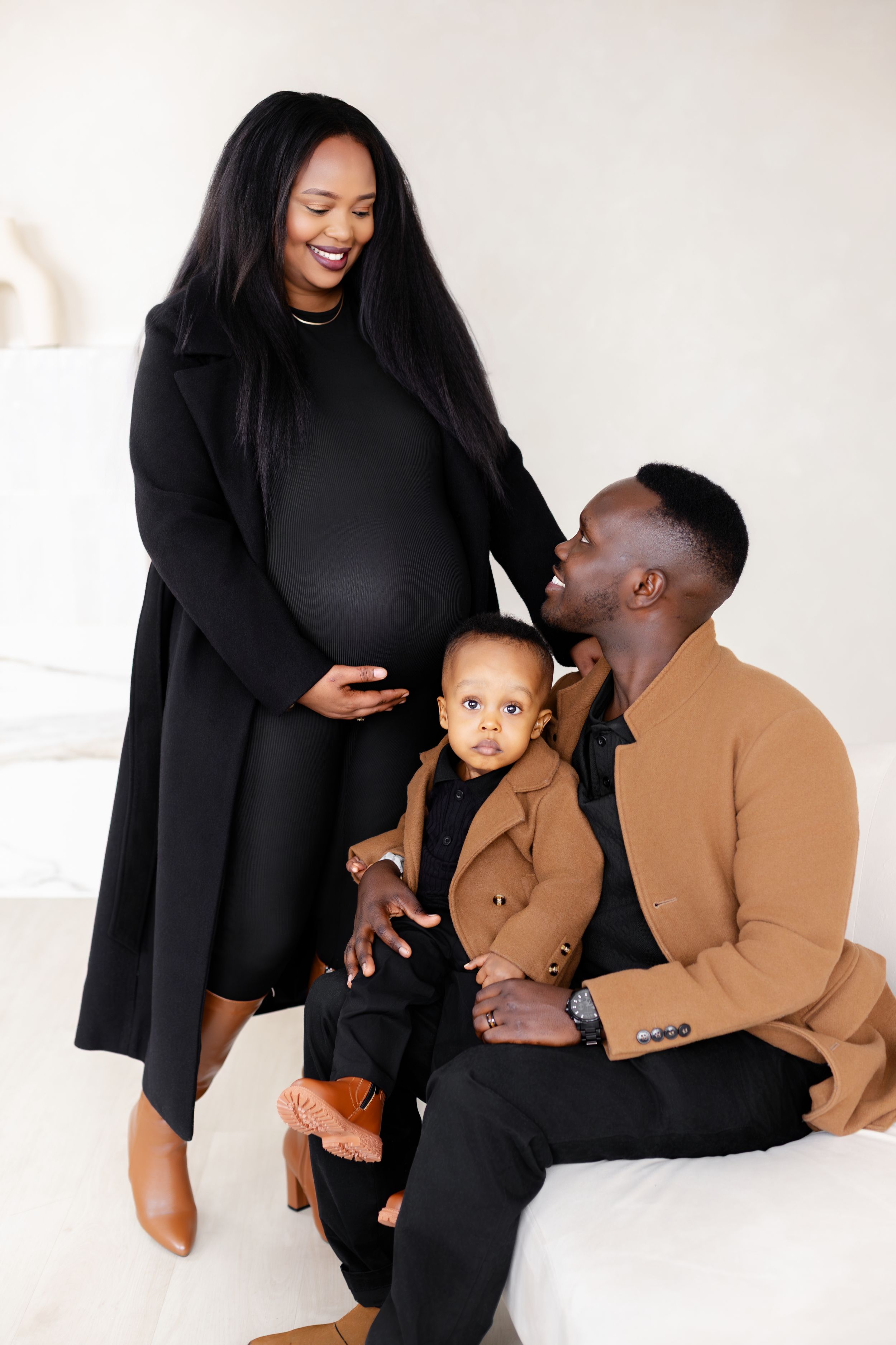 A family of three poses together in coordinated black and brown outfits against a white studio backdrop.