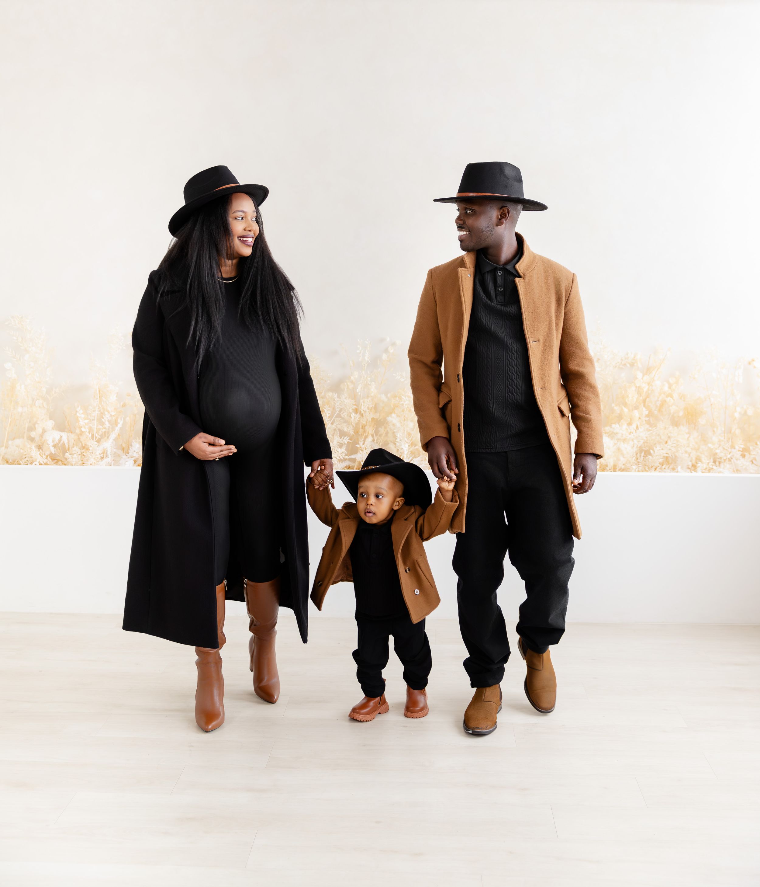 A couple in matching black outfits and brown boots poses together in a minimalist white studio setting.