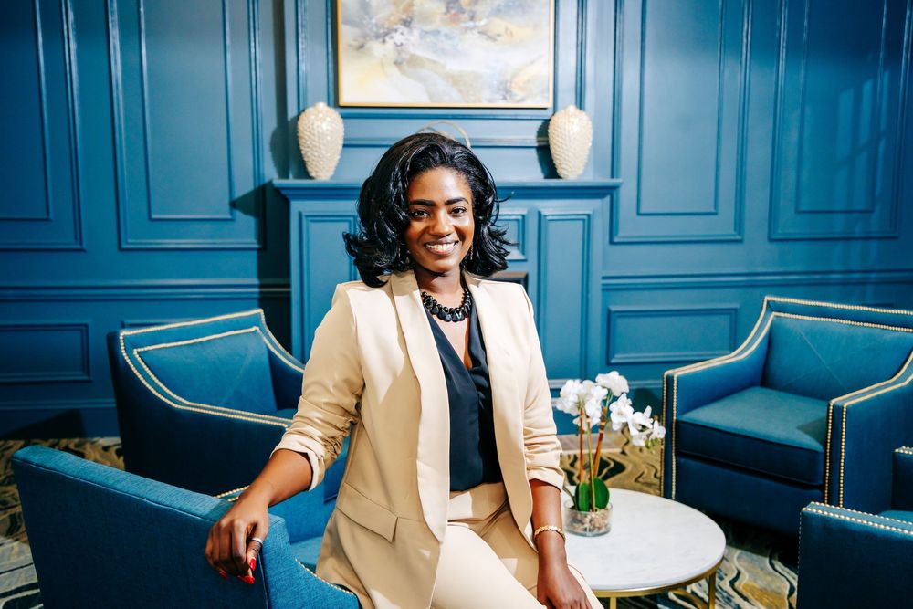 Professional woman in beige blazer smiling confidently in elegant blue-paneled room with teal chairs.