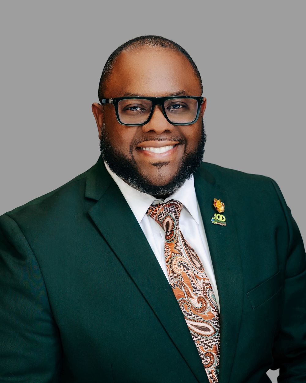 Professional man in green blazer and patterned tie smiling confidently against gray background.