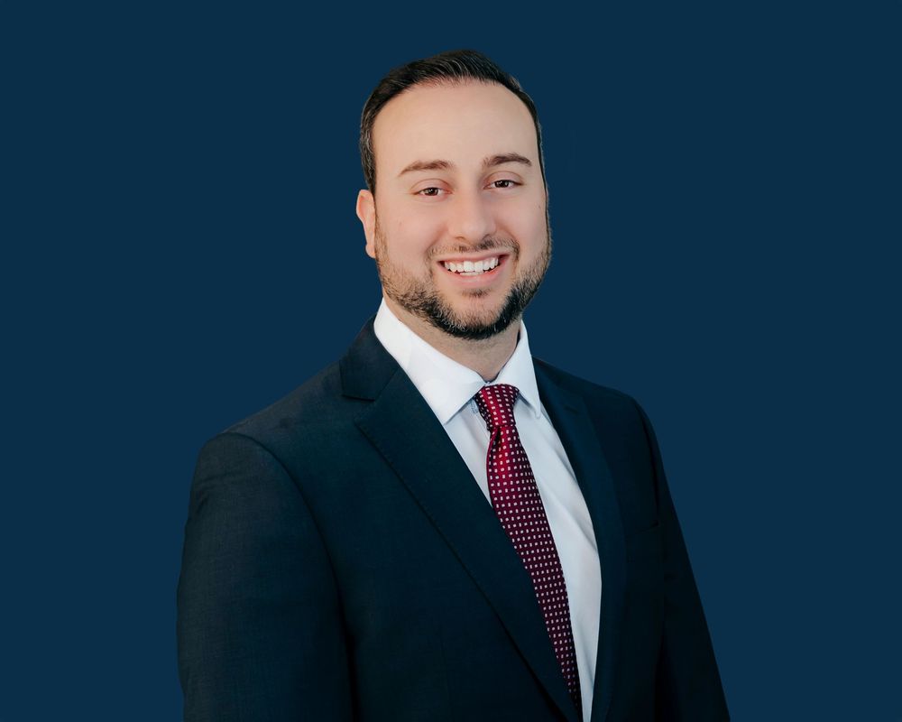Professional headshot of a smiling young businessman in a navy suit and red tie against a dark blue background.